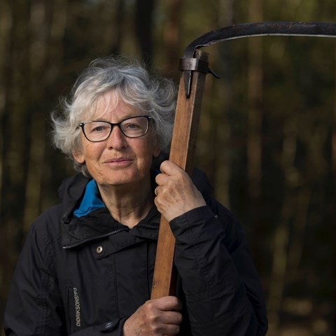 Kvinna med grått hår och blå jacka stor i skogen och lutar sig mot en lie. Foto