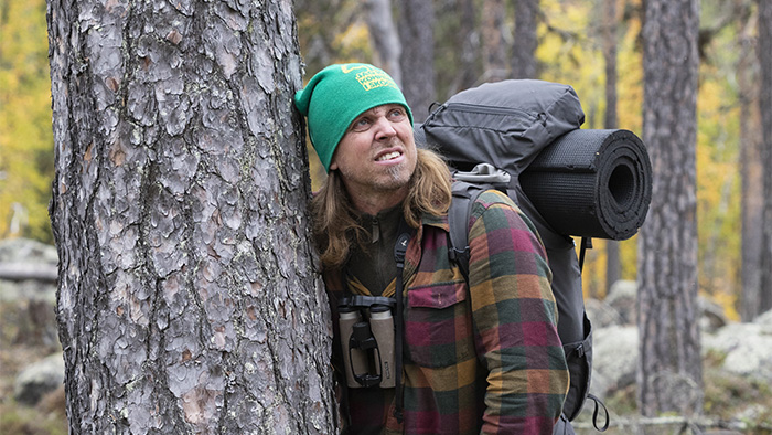 Man i friluftskläder och med campingutrustning på ryggen står lutad mot träd i skogen. Foto