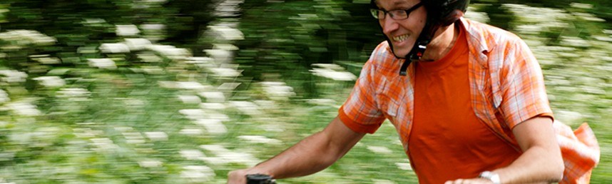 Leende man i orange kortärmad skjorta och t-shirt cyklar i fart med grönska i bakgrunden. Foto