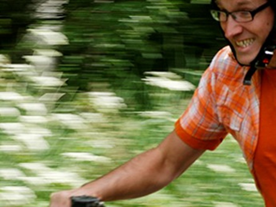 Leende man i orange kortärmad skjorta och t-shirt cyklar i fart med grönska i bakgrunden. Foto